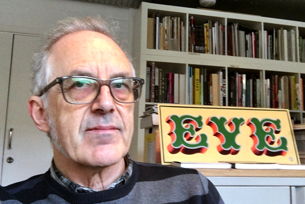 Headshot of man in glasses in front of book shelf> Handpianted sign reads ‘Eye’.